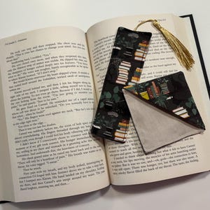 May include: A black and green fabric bookmark with a tassel. The bookmark features a pattern of stacked books and plants. The bookmark is folded in half and placed on top of an open book.