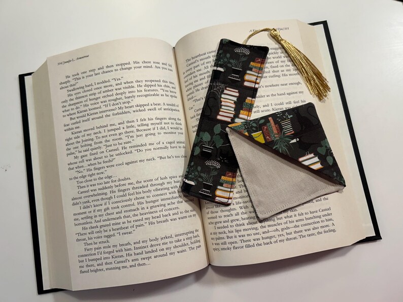 May include: A black and green fabric bookmark with a tassel. The bookmark features a pattern of stacked books and plants. The bookmark is folded in half and placed on top of an open book.