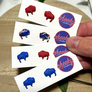 May include: Four pairs of stud earrings shaped like bison, displayed on white cards with the text cheeky creations. The earrings feature solid red, solid blue, and marbled red, white, and blue designs with gold fleck accents on the marbled pair.