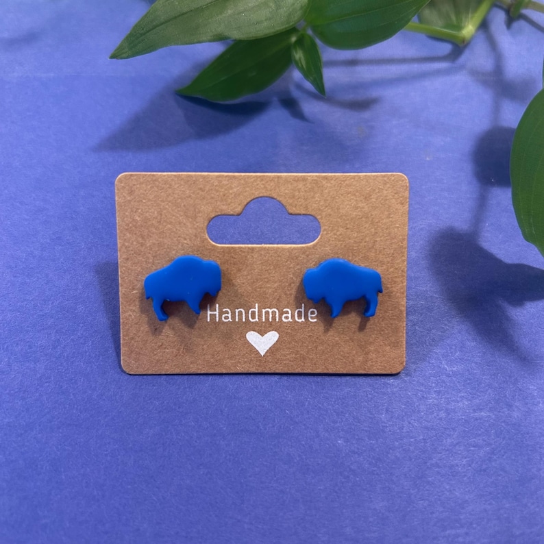 May include: A pair of blue buffalo-shaped stud earrings displayed on a brown card with the word "Handmade" and a white heart. The earrings are a vibrant blue color and are presented on a blue background.