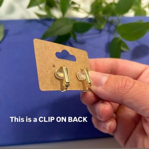 May include: A pair of gold-colored clip-on earrings displayed on a brown card with a blue background. The text "This is a CLIP ON BACK" is visible on the card. The earrings have a simple, elegant design.