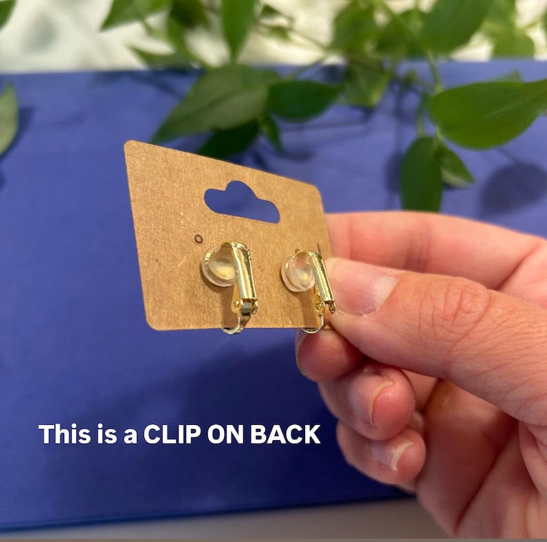 May include: A pair of gold-colored clip-on earrings displayed on a brown card with a blue background. The text "This is a CLIP ON BACK" is visible on the card. The earrings have a simple, elegant design.