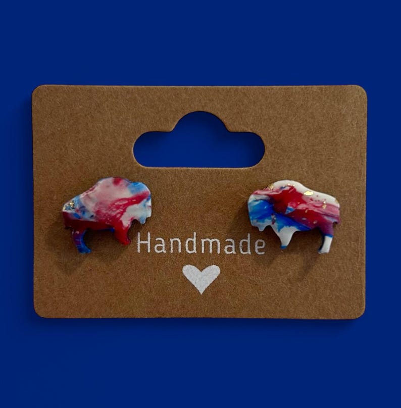 May include: A pair of handmade buffalo-shaped earrings. The earrings are red, white, and blue with gold accents. They are displayed on a brown card with the word "Handmade" and a heart symbol.