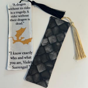 May include: Two fabric bookmarks with black tassels. One bookmark features a gold dragon and the quote "A dragon without its rider is a tragedy. A rider without their dragon is dead." The other bookmark has a black and gray dragon scale pattern and the quote "I know exactly who and what you are, Violet Sorrengail."