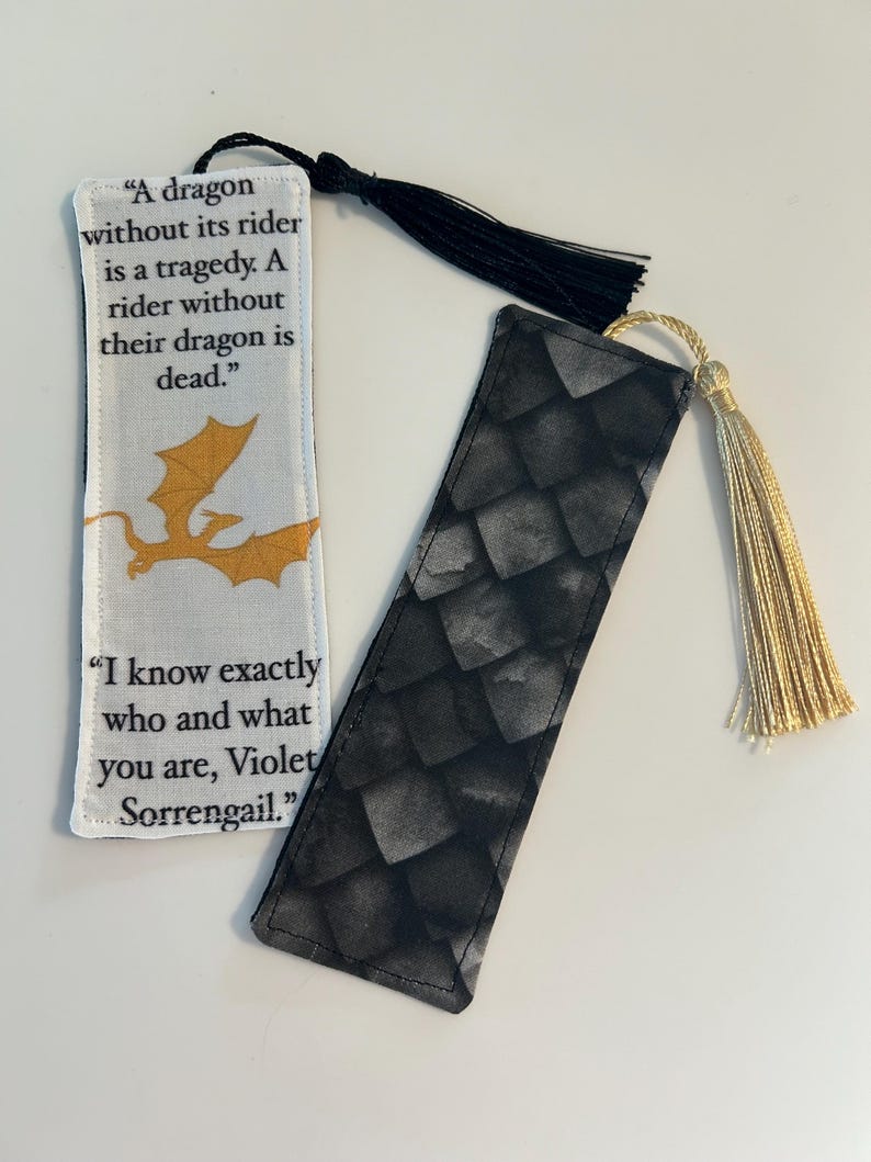 May include: Two fabric bookmarks with black tassels. One bookmark features a gold dragon and the quote "A dragon without its rider is a tragedy. A rider without their dragon is dead." The other bookmark has a black and gray dragon scale pattern and the quote "I know exactly who and what you are, Violet Sorrengail."