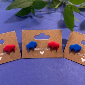 May include: Three pairs of handmade stud earrings on brown cardboard displays. Each earring is shaped like a buffalo. Two pairs have red buffaloes, and one pair has blue buffaloes. The displays have the word "Handmade" and a heart.