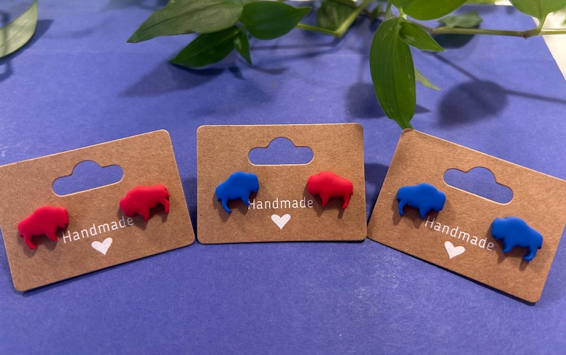 May include: Three pairs of handmade stud earrings on brown cardboard displays. Each earring is shaped like a buffalo. Two pairs have red buffaloes, and one pair has blue buffaloes. The displays have the word "Handmade" and a heart.