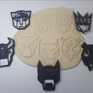 Könnte beinhalten: Ein Set aus neun schwarzen Plastik-Ausstechern mit verschiedenen Transformers-Charakteren. Die Ausstecher sind auf einem Blatt weißen Teigs angeordnet.