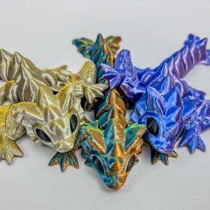 Könnte beinhalten: Vier kleine, 3D-gedruckte Drachenfiguren in verschiedenen Farben: Silber, Gold, Blau und Grün. Die Drachen sind spielerisch ineinander verschlungen.