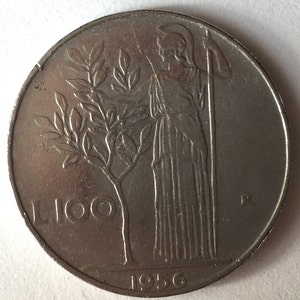 May include: A silver Italian coin with the year 1956 and the denomination "L. 100" on the front. The coin features a woman standing with a staff and a tree with leaves behind her.