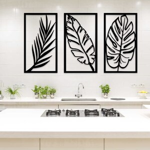 May include: Three black metal wall art pieces, each framed in a rectangular black border, depicting stylized tropical leaves. The artwork hangs above a white kitchen countertop with a stainless steel faucet and a gas cooktop.
