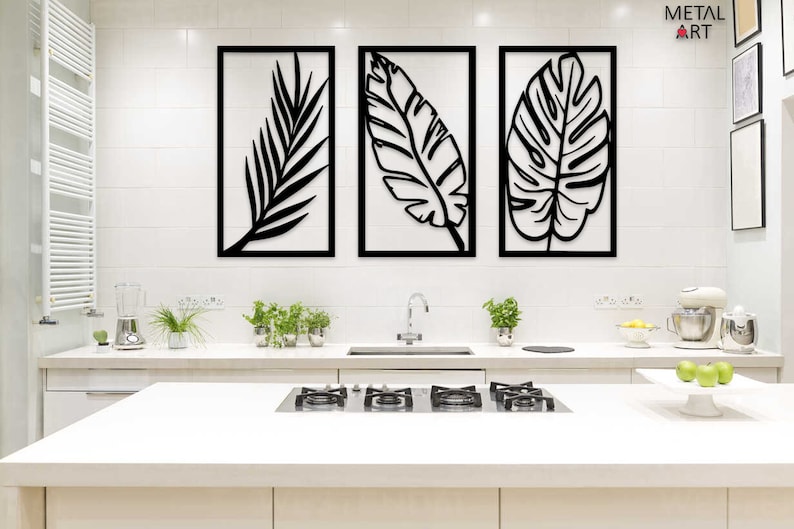 May include: Three black metal wall art pieces, each framed in a rectangular black border, depicting stylized tropical leaves. The artwork hangs above a white kitchen countertop with a stainless steel faucet and a gas cooktop.