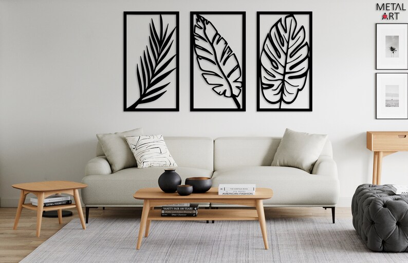 May include: Three black metal wall art pieces with leaf designs in rectangular frames. The artwork is displayed above a light-colored sofa and wooden coffee table, creating a modern, minimalist aesthetic. The room is decorated with neutral tones.