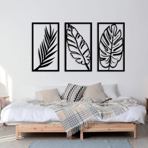 May include: Three black metal leaf wall art pieces in rectangular frames. The artwork is displayed above a bed with a wooden frame, white bedding, and decorative pillows. The room has a neutral color palette.