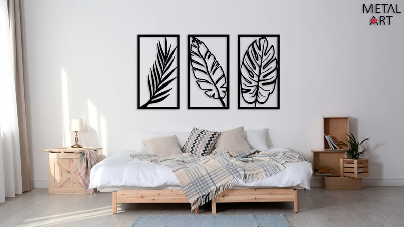 May include: Three black metal leaf wall art pieces in rectangular frames. The artwork is displayed above a bed with a wooden frame, white bedding, and decorative pillows. The room has a neutral color palette.