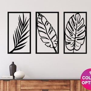 May include: Three framed wall art pieces featuring black leaf silhouettes against a white background. The leaves are in different shapes and sizes. The text "12 COLOUR OPTIONS" is in a pink circle. The art is displayed above a wooden cabinet.