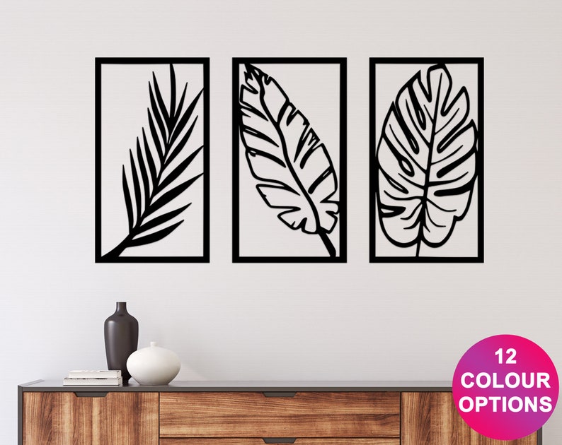 May include: Three framed wall art pieces featuring black leaf silhouettes against a white background. The leaves are in different shapes and sizes. The text "12 COLOUR OPTIONS" is in a pink circle. The art is displayed above a wooden cabinet.