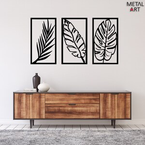 May include: Three black metal leaf wall art pieces in rectangular frames. The leaves have intricate vein details and are set against a white background. The artwork is displayed above a wooden cabinet with a dark gray top.