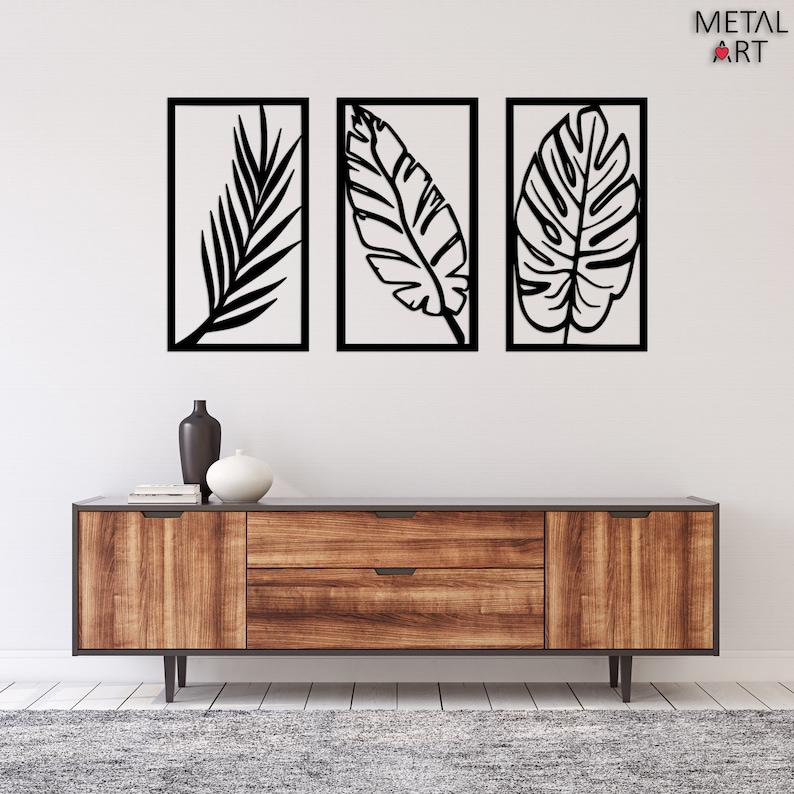 May include: Three black metal leaf wall art pieces in rectangular frames. The leaves have intricate vein details and are set against a white background. The artwork is displayed above a wooden cabinet with a dark gray top.