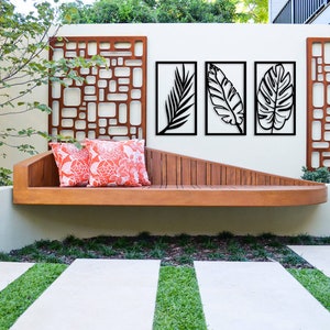 May include: Outdoor scene featuring a wooden bench with two orange floral pillows. Above the bench are three framed black leaf art pieces and two decorative metal panels. The wall is painted a light beige color, and the ground has a stone walkway with grass.