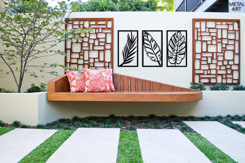 May include: Outdoor scene featuring a wooden bench with two orange floral pillows. Above the bench are three framed black leaf art pieces and two decorative metal panels. The wall is painted a light beige color, and the ground has a stone walkway with grass.