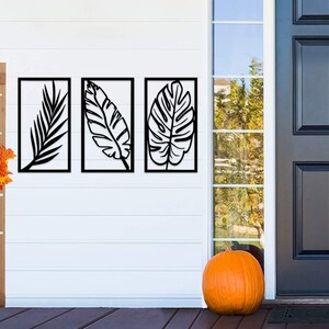 May include: Three black metal leaf wall art panels. Each panel features a different leaf design within a rectangular frame. A wooden sign with the word "HOME" and a fall wreath are to the left. An orange pumpkin sits on the porch.