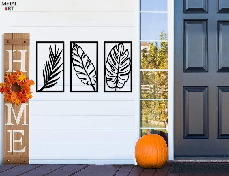 May include: Three black metal leaf wall art panels. Each panel features a different leaf design within a rectangular frame. A wooden sign with the word "HOME" and a fall wreath are to the left. An orange pumpkin sits on the porch.