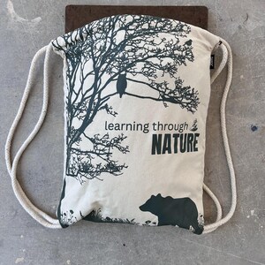 Puede incluir: Una mochila con cordón de color crema con un diseño de árbol y oso verde oscuro. El texto "learning through NATURE" está impreso en la parte delantera. La bolsa tiene correas de cuerda blanca y está colocada sobre una superficie marrón.