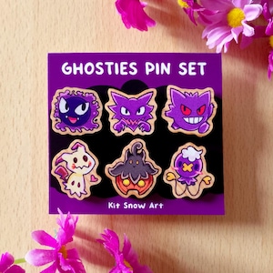 May include: A purple card with the text "GHOSTIES PIN SET" displays six colorful character pins. The pins feature various cartoon ghost designs in purple, black, and cream colors. The text "Kit Snow Art" is at the bottom.