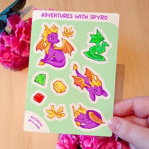 May include: A sticker sheet with the text "ADVENTURES WITH SPYRO" features cartoon images of a purple dragon, green dragon, gems, and a butterfly. The sticker sheet has a light green background and the website KitSnowArt.com is printed on the bottom left.
