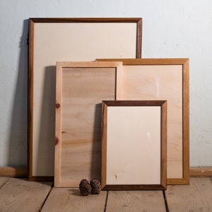 Könnte beinhalten: Vier Holzrahmen in verschiedenen Größen und Farben, übereinander gestapelt. Die Rahmen sind leer und haben einen hellbraunen Hintergrund. Die Rahmen stehen auf einem Holzboden.