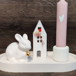 Könnte beinhalten: Weiße Keramik-Hasenfigur mit einer goldenen Glocke, ein weißes Holzhaus mit rosa Blumen und eine rosa Kerze auf einem weißen ovalen Tablett.