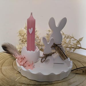 Könnte beinhalten: Weißer Kerzenhalter aus Keramik in Form eines Osterhasen mit einer rosa Kerze und einer kleinen Feder. Der Halter hat einen gewellten Rand und ein kleines Holzschild mit der Aufschrift "Ostergruss".