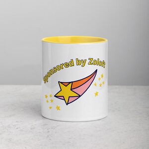 Puede incluir: Taza de cer&aacute;mica blanca con un borde amarillo. La taza tiene una estrella fugaz rosa y naranja con una estrella amarilla en la parte superior. El texto "Sponsored by Zoloft" est&aacute; escrito en un c&iacute;rculo amarillo alrededor de la estrella.