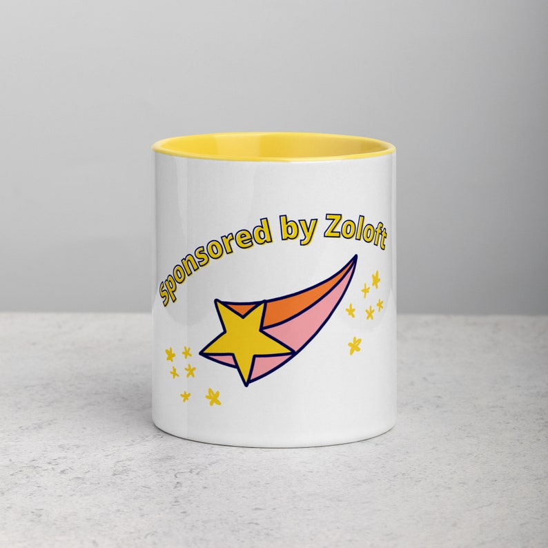 Puede incluir: Taza de cer&aacute;mica blanca con un borde amarillo. La taza tiene una estrella fugaz rosa y naranja con una estrella amarilla en la parte superior. El texto "Sponsored by Zoloft" est&aacute; escrito en un c&iacute;rculo amarillo alrededor de la estrella.