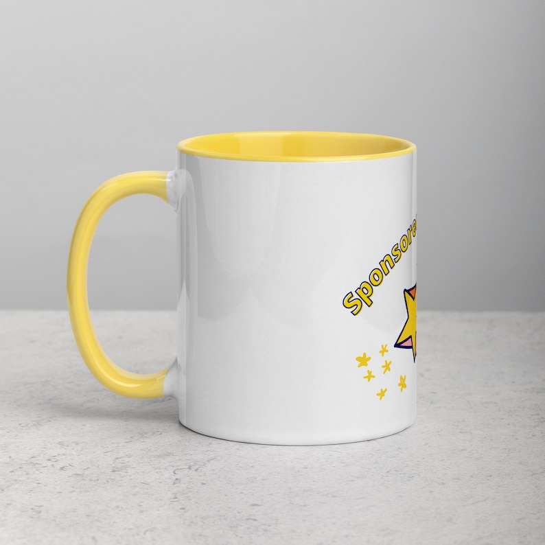 Puede incluir: Una taza de cer&aacute;mica blanca con un borde y un asa amarillos. La taza tiene una estrella amarilla y el texto "Sponsored by" impreso en ella.