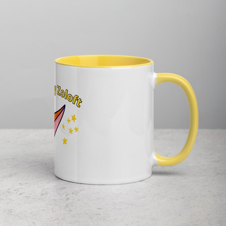Puede incluir: Taza de cer&aacute;mica blanca con borde y asa amarillos. La taza tiene un cohete de dibujos animados rosa y naranja con el texto "Happy Zoloft" en amarillo.