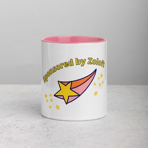 Puede incluir: Taza de cer&aacute;mica blanca con un borde rosa. La taza tiene una estrella amarilla que se dispara a trav&eacute;s del frente con el texto "Sponsored by Zoloft" en un arco amarillo sobre la estrella.