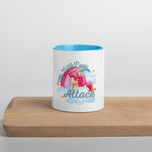 Peut inclure: Une tasse en céramique blanche avec un bord bleu clair. La tasse représente une licorne de dessin animé avec une crinière et une queue roses, entourée de nuages et d'étoiles. Le texte "My Little Panic Attack" est écrit en lettres turquoise.