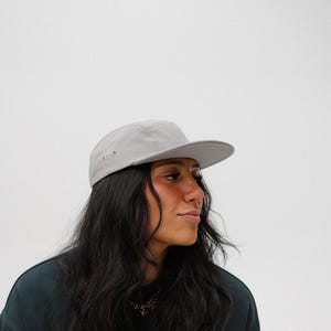 Puede incluir: Una persona con una gorra gris de 5 paneles y una sudadera negra. La persona tiene el pelo largo y oscuro.