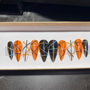May include: Set of ten stiletto-shaped press-on nails. The nails alternate between orange, white, and black bases, each adorned with a grid pattern of black, gold, and white lines. The nails are displayed in a white box.