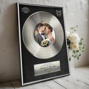 Könnte beinhalten: Eine silberne Schallplatten-Auszeichnung mit schwarzem Rahmen und einem Foto eines Paares in der Mitte. Die Auszeichnung ist für 25 Jahre Ehe und lautet "Auszeichnung für 25 glückliche Jahre Sandra & Christian 24.01.1999".