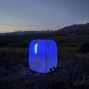 Könnte beinhalten: Ein blau-weißes Pop-up-Zelt, das mit einem vertikalen blauen Lichtstreifen beleuchtet wird. Das Zelt steht im Freien auf einer Wiese mit Bergen im Hintergrund. Ein zweites Zelt ist in der Ferne zu sehen.