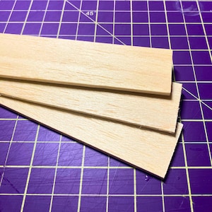 May include: Three pieces of unfinished, light brown wood, stacked on top of each other. The wood is smooth and has a natural grain. The wood is on a purple cutting mat.