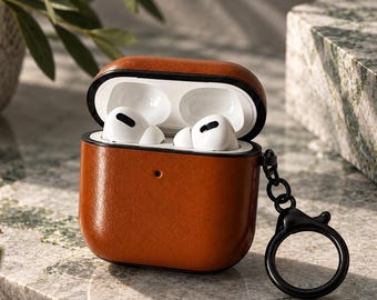 May include: A cognac-colored leather protective case for wireless earbuds, featuring a black interior lining and a black metal keychain ring attached to the side. The case is open, revealing two white wireless earbuds stored inside the charging compartment.