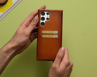 Puede incluir: Una funda de teléfono de cuero marrón con tarjetero. La funda tiene forma rectangular con esquinas redondeadas y un recorte para la cámara. Dos tarjetas de crédito son visibles en las ranuras para tarjetas. La funda está sobre un fondo verde.