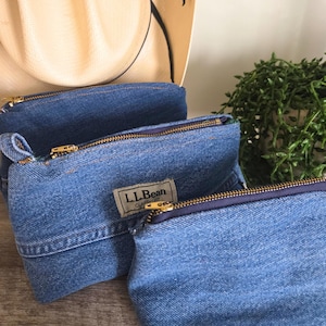 May include: Three denim pouches with gold zippers and a small L.L.Bean label. The pouches are made from blue denim fabric and have a classic fit. A straw hat is visible in the background.