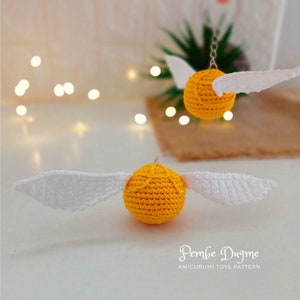 May include: Two crocheted golden snitch toys with white wings. The snitch on the left is sitting on a white surface, while the snitch on the right is hanging from a chain.