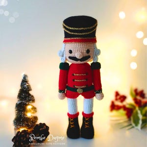 May include: A crocheted nutcracker doll in a soldier's uniform. The doll has a black hat with gold trim, a red jacket, white trousers, and black boots. The doll has a white moustache and curly hair.