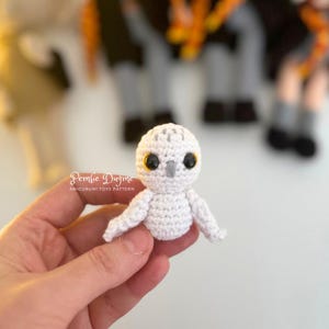 May include: A small, white crocheted owl with black eyes and a gray beak is held in a hand. The owl has small wings and is part of an amigurumi toy pattern. The text "Pembe Dugme AMIGURUMI TOYS PATTERN" is visible.
